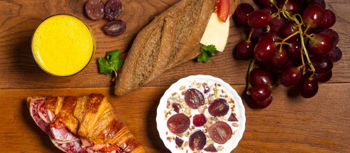 Flat lay of croissant, baguette, grapes, yogurt, and juice on a wooden table.