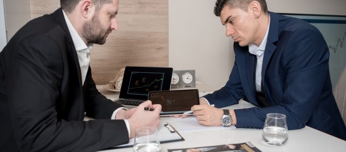 Two businessmen in suits discussing cryptocurrency trends using digital devices in an office setting.